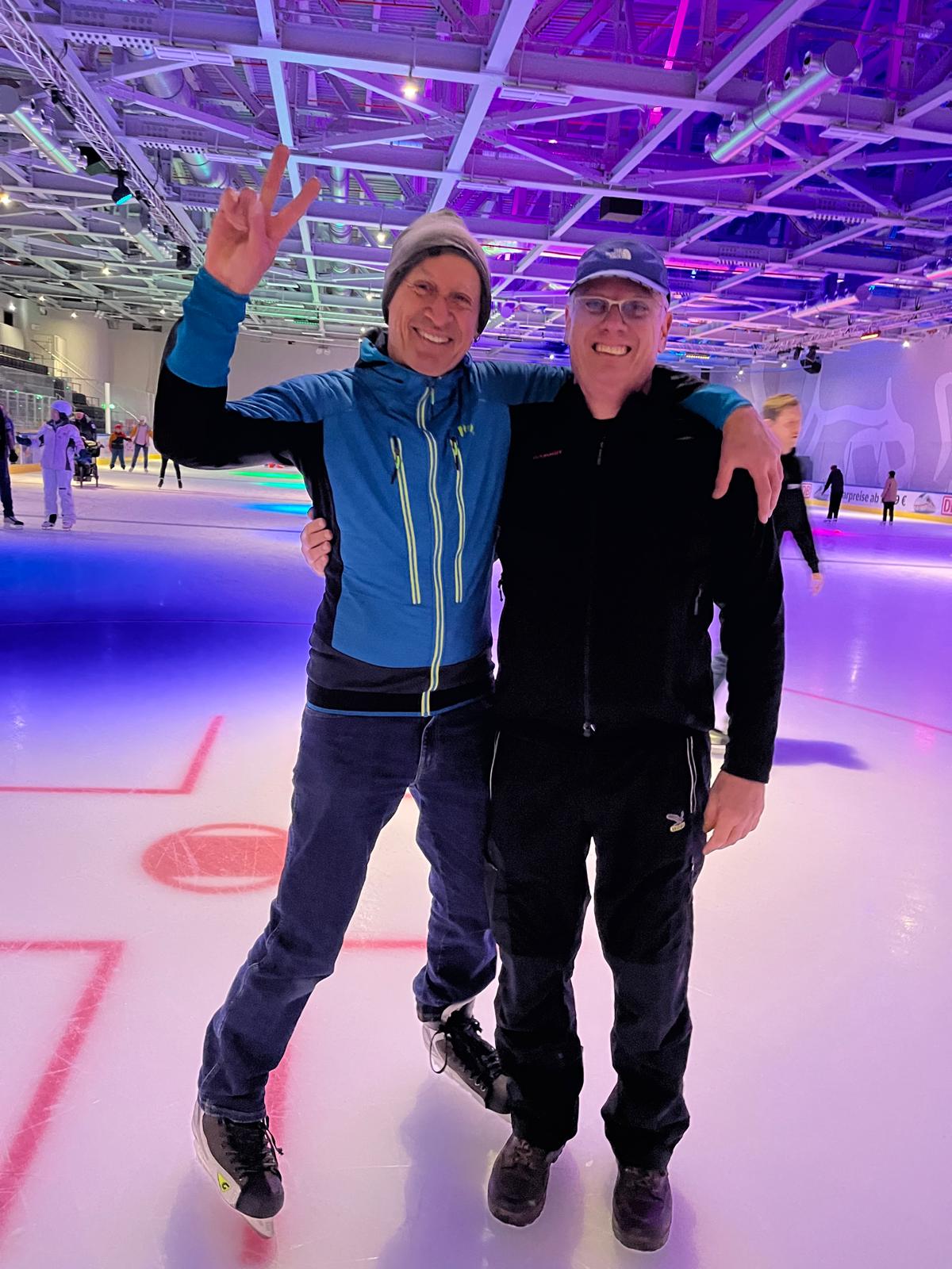 Zwei lächelnde Männer stehen gemeinsam auf einer beleuchteten Eislauffläche. Der Mann links hebt die Hand zu einem Victory-Zeichen, beide tragen Winterkleidung und stehen Arm in Arm. Im Hintergrund sind weitere Schlittschuhläufer und farbiges Deckenlicht zu sehen.