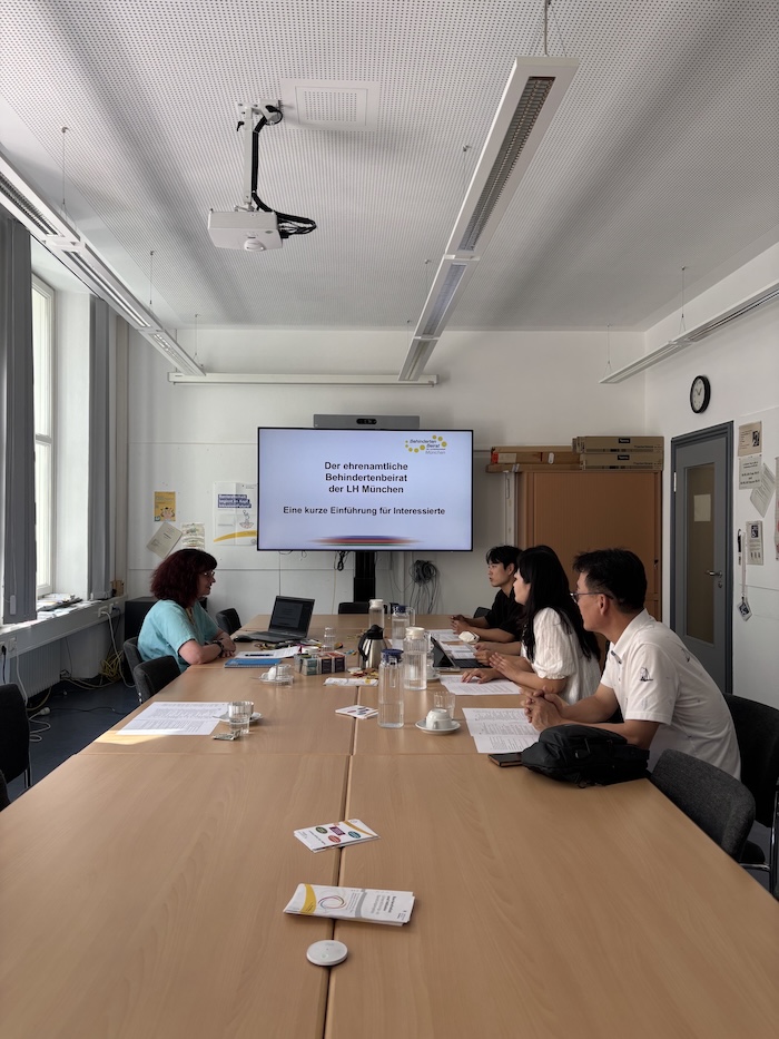 Am Tisch sitzen links Nadja Rackwitz und rechts die Delegation aus Korea mit 4 Personen. Es ist ein Monitor mit einer Präsentation zum Behindertenbeirat zu sehen.