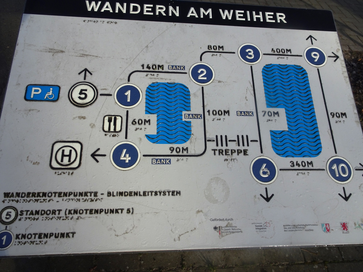 Das Bild zeigt eine tastbare Wanderkarte mit dem Titel "WANDERN AM WEIHER", die speziell für blinde und sehbehinderte Menschen entwickelt wurde. Dies ist an der Kombination aus Großdruck, Brailleschrift (Blindenschrift) und erhabenen, tastbaren Linien und Symbolen erkennbar